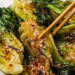 Chopsticks lifting up a piece of baby bok choy with garlic and ginger from a serving platter with a text overlay on the photo.