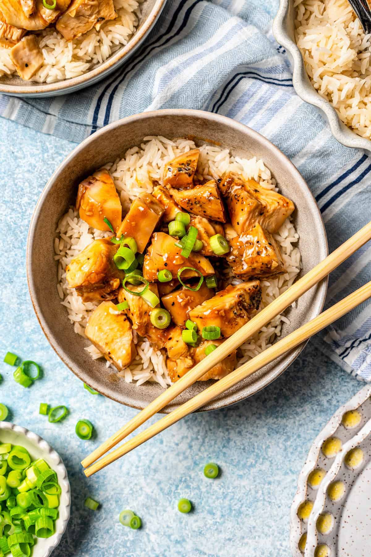Close up of a bowl of bourbon chicken over rice with chopped green onions on top and to the side and a pair of chopsticks resting on the bowl.