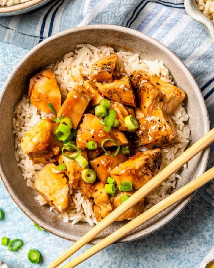 Square cut of a bowl of bourbon chicken over rice with chopped green onions on top and to the side and a pair of chopsticks resting on the bowl.