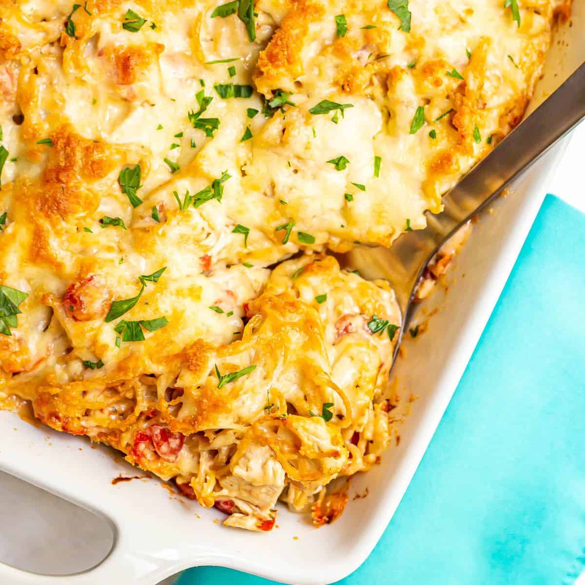 Close up of a serving spoon scooping up some baked chicken spaghetti from a large white casserole dish.