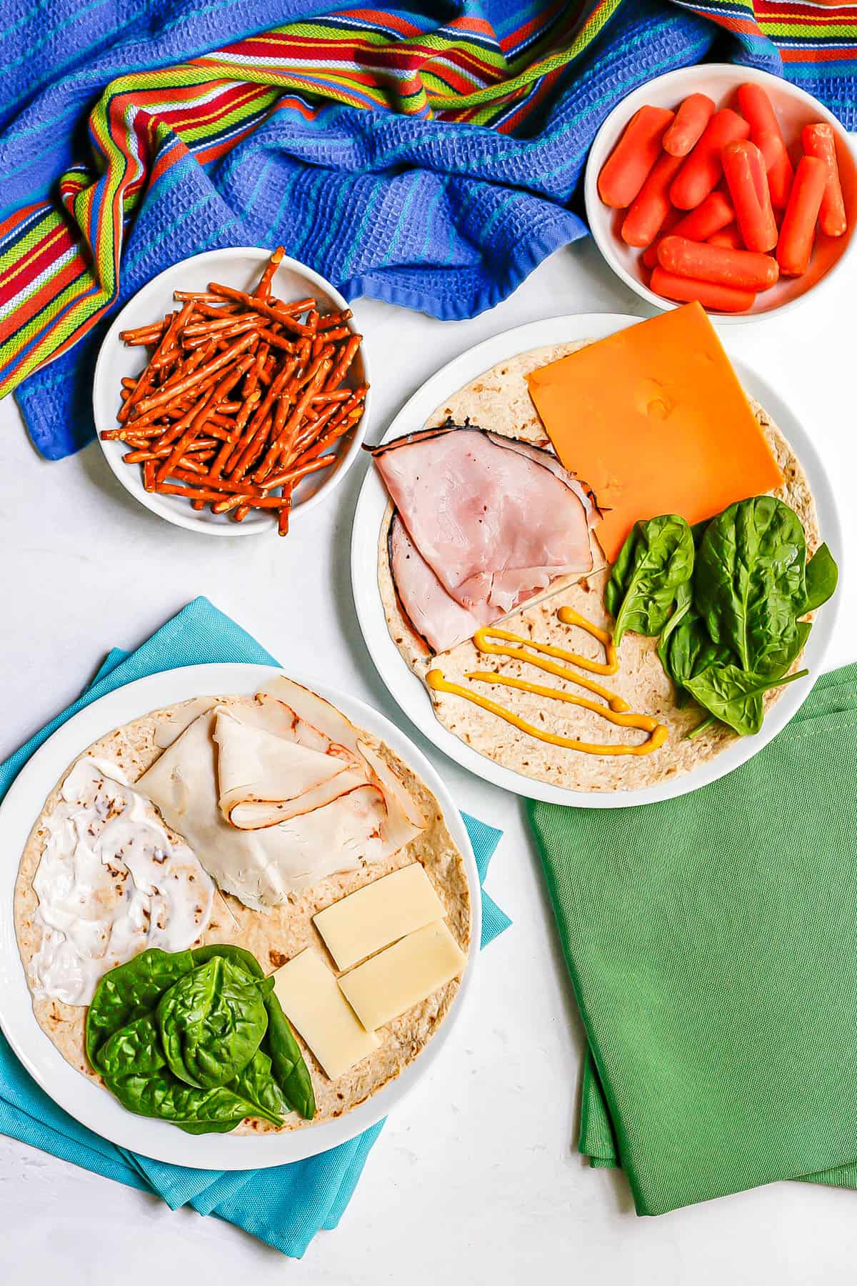 Folded wraps laid out on two white plates, one with a tortilla topped with ham and cheddar and spinach and mustard, the other with turkey and cheese and spinach and mayo.