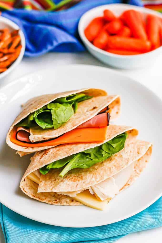 A ham and cheese folded wrap atop a turkey and cheese folded wrap on a white plate with pretzels and baby carrots in the background.