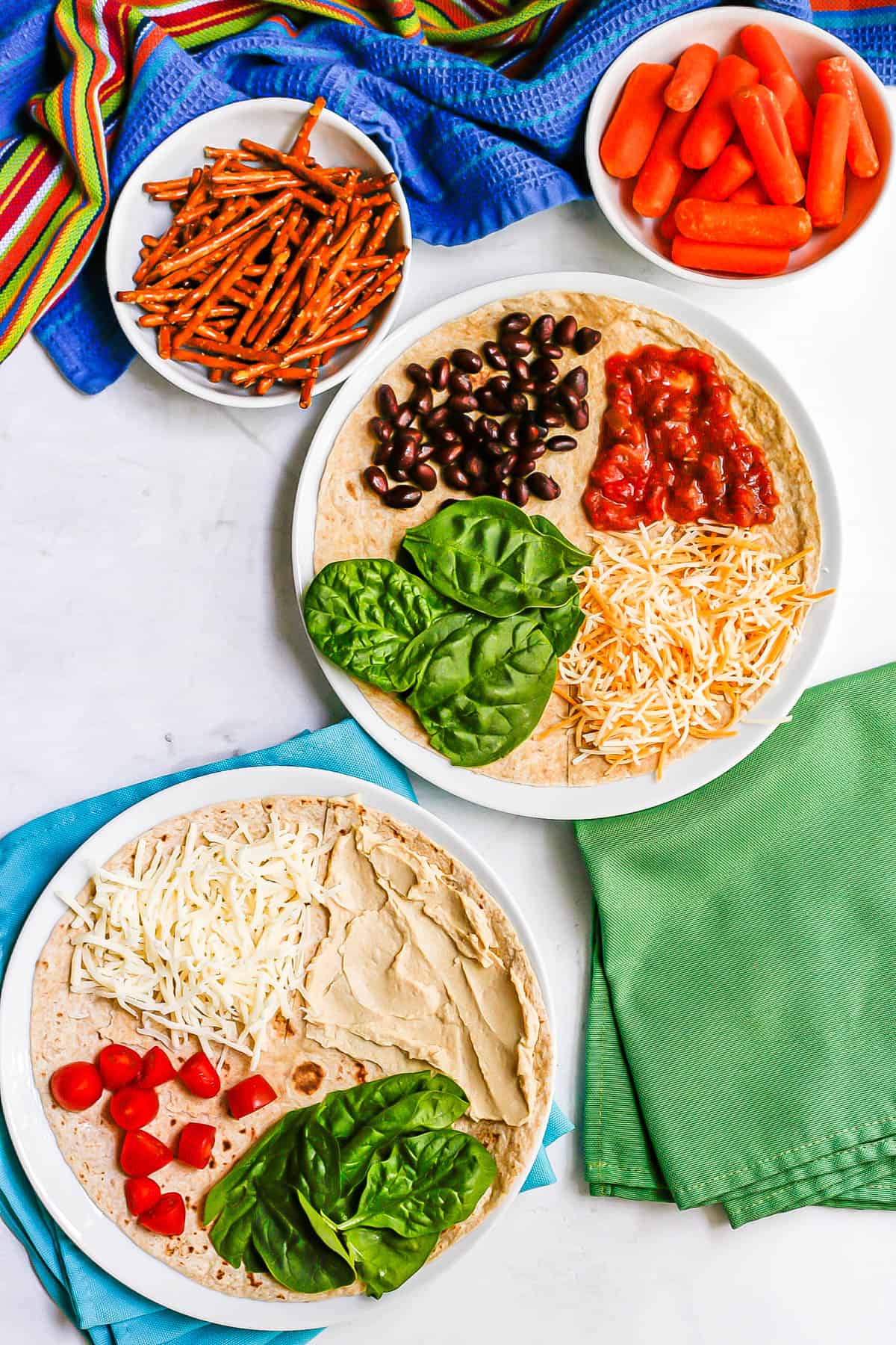 Two vegetarian folded wraps on white plates, one with black beans, cheese, salsa and spinach and the other with hummus, cheese, spinach and tomatoes.