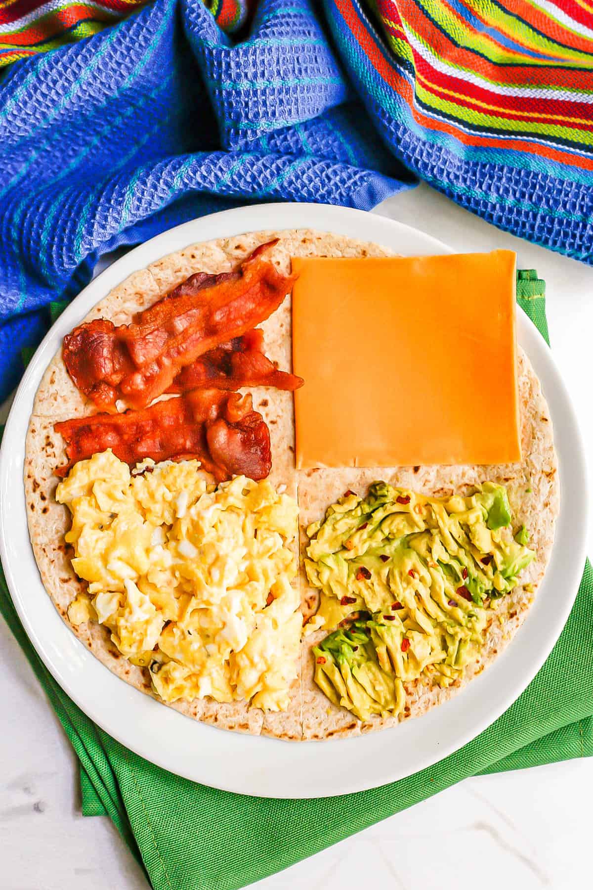 A breakfast folded wrap tortilla on a plate with quadrants of scrambled egg, bacon, cheddar cheese and mashed avocado.