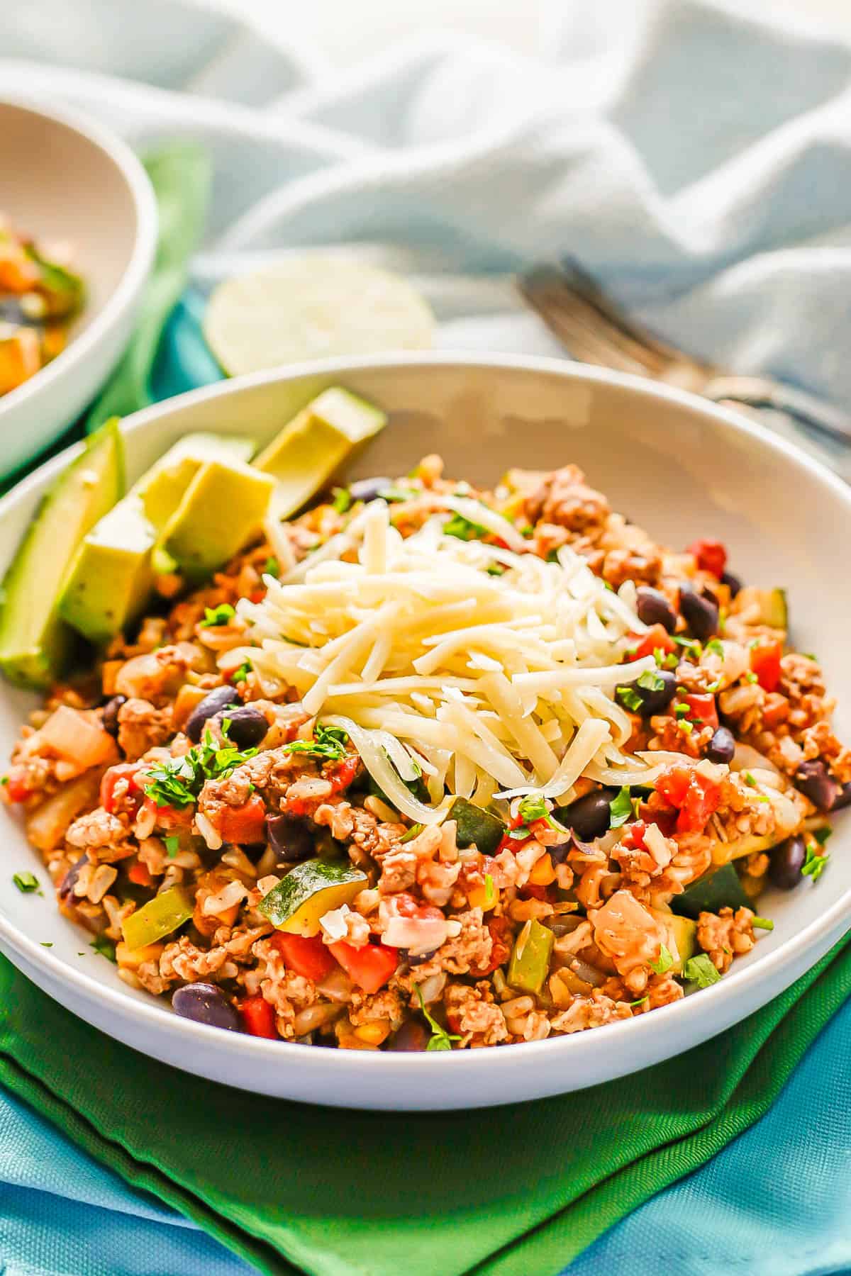 A white bowl with a serving of a ground turkey, rice and veggie skillet mix with shredded cheese on top and avocado slices to the side.
