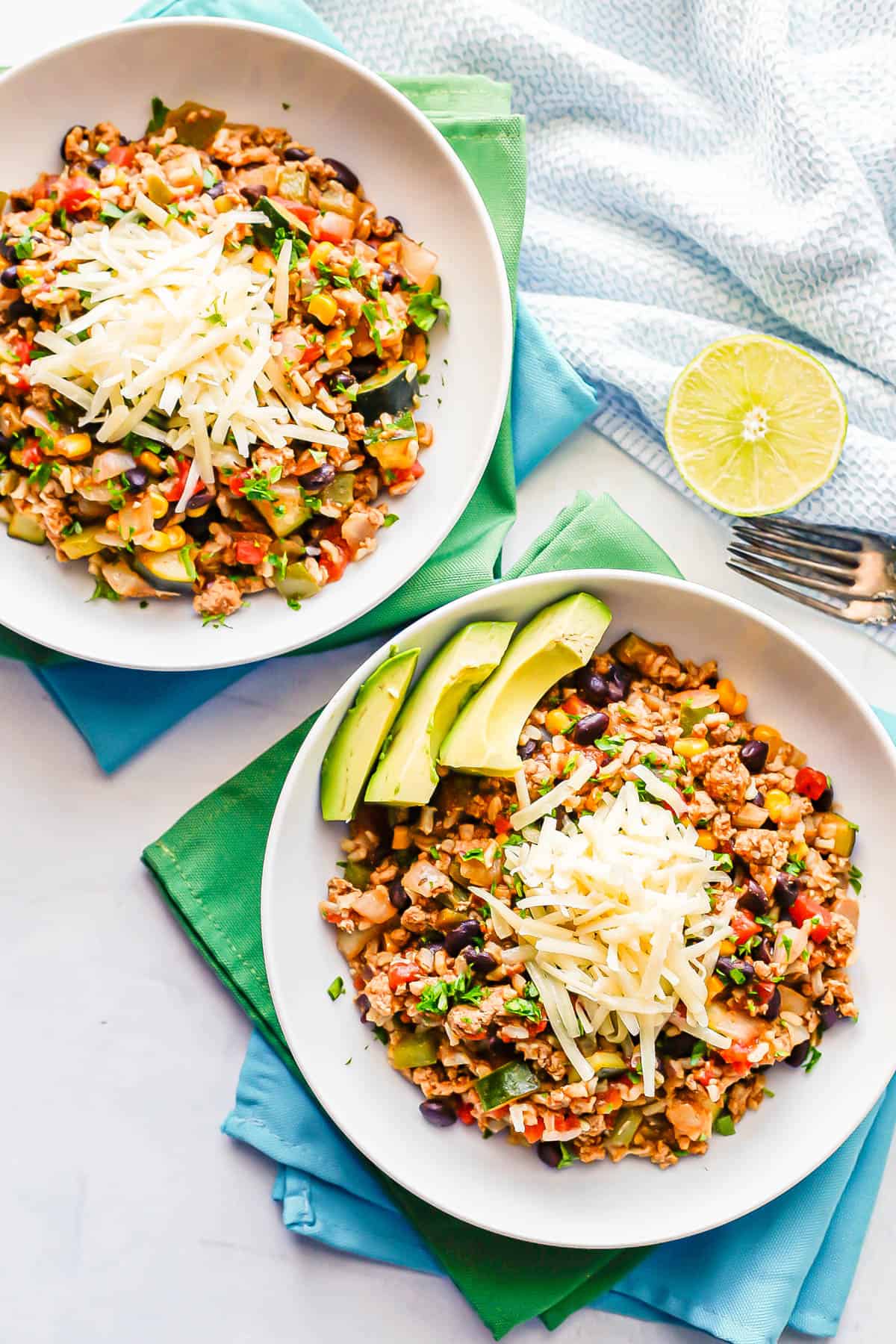 Two low white bowls filled with a turkey rice and veggie mixture topped with shredded cheese and avocado slices.