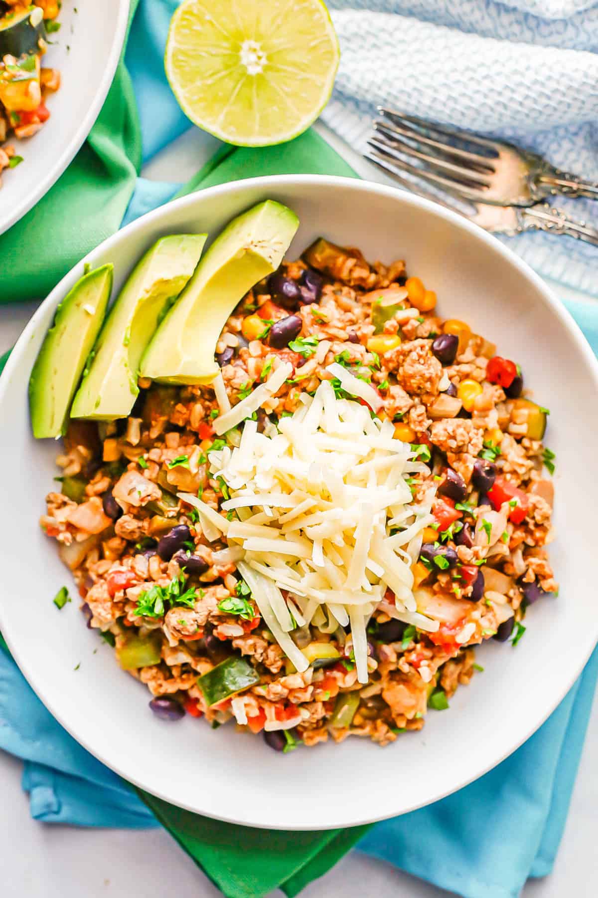 Close up of a serving of a ground turkey, rice and veggie skillet mix in a white bowl with shredded cheese on top and avocado slices to the side.