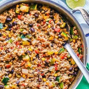 Close up of a silver serving spoon resting in a large pot with a mixture of ground turkey, rice, beans and veggies.