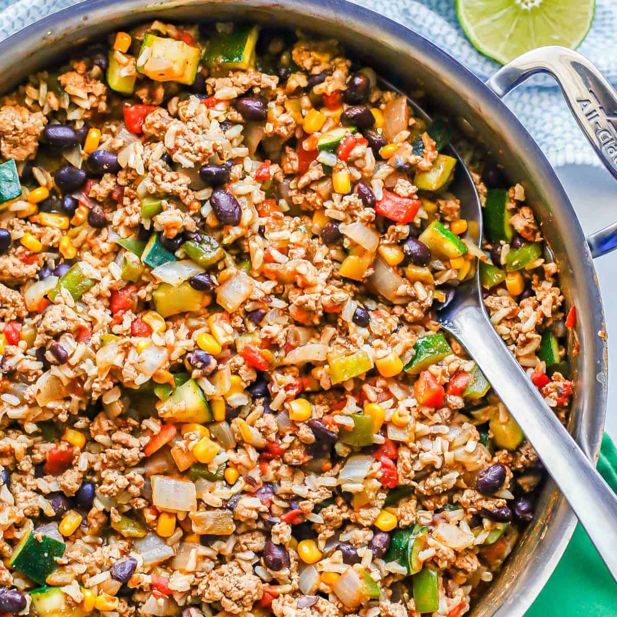 Close up of a silver serving spoon resting in a large pot with a mixture of ground turkey, rice, beans and veggies.