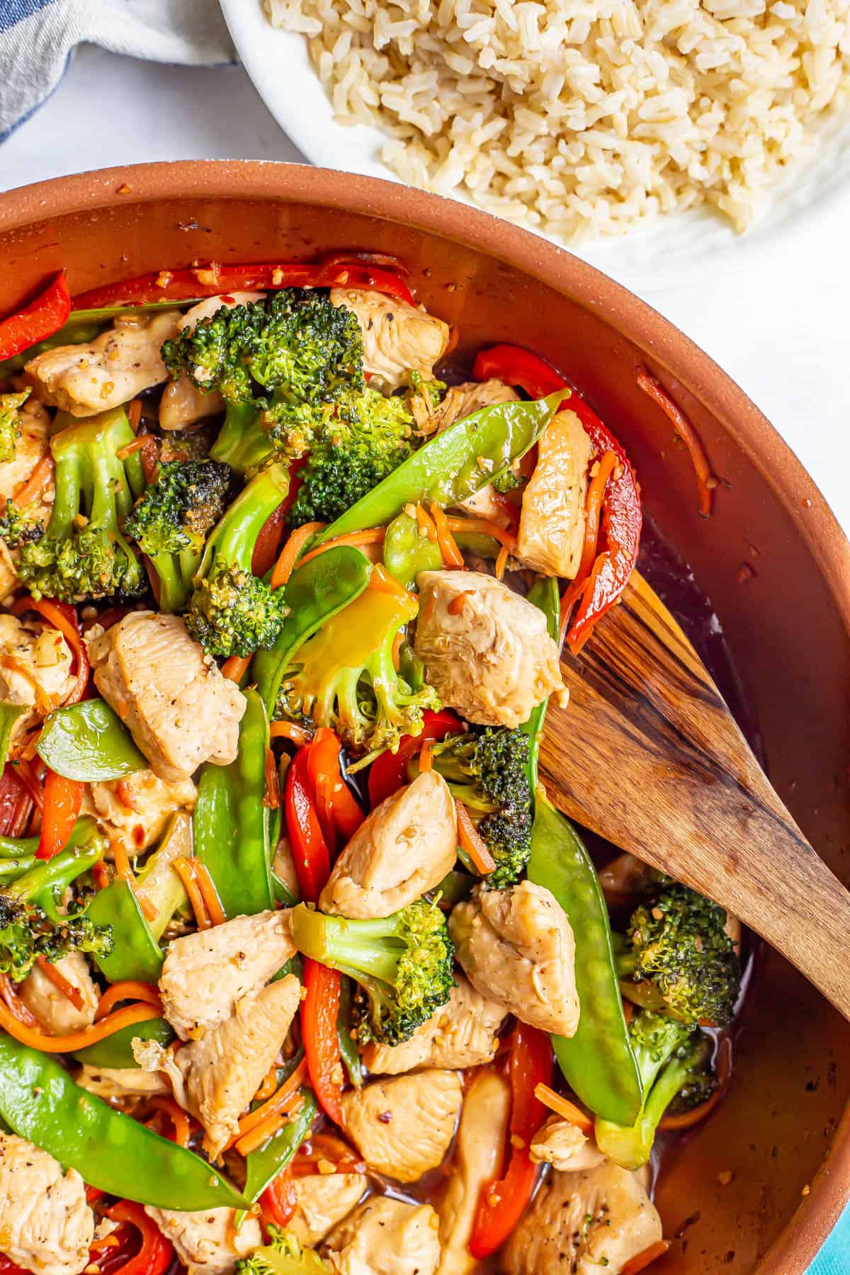 Close up of chicken and veggie stir fry with a honey garlic sauce in a large skillet with a wooden spoon resting in it.