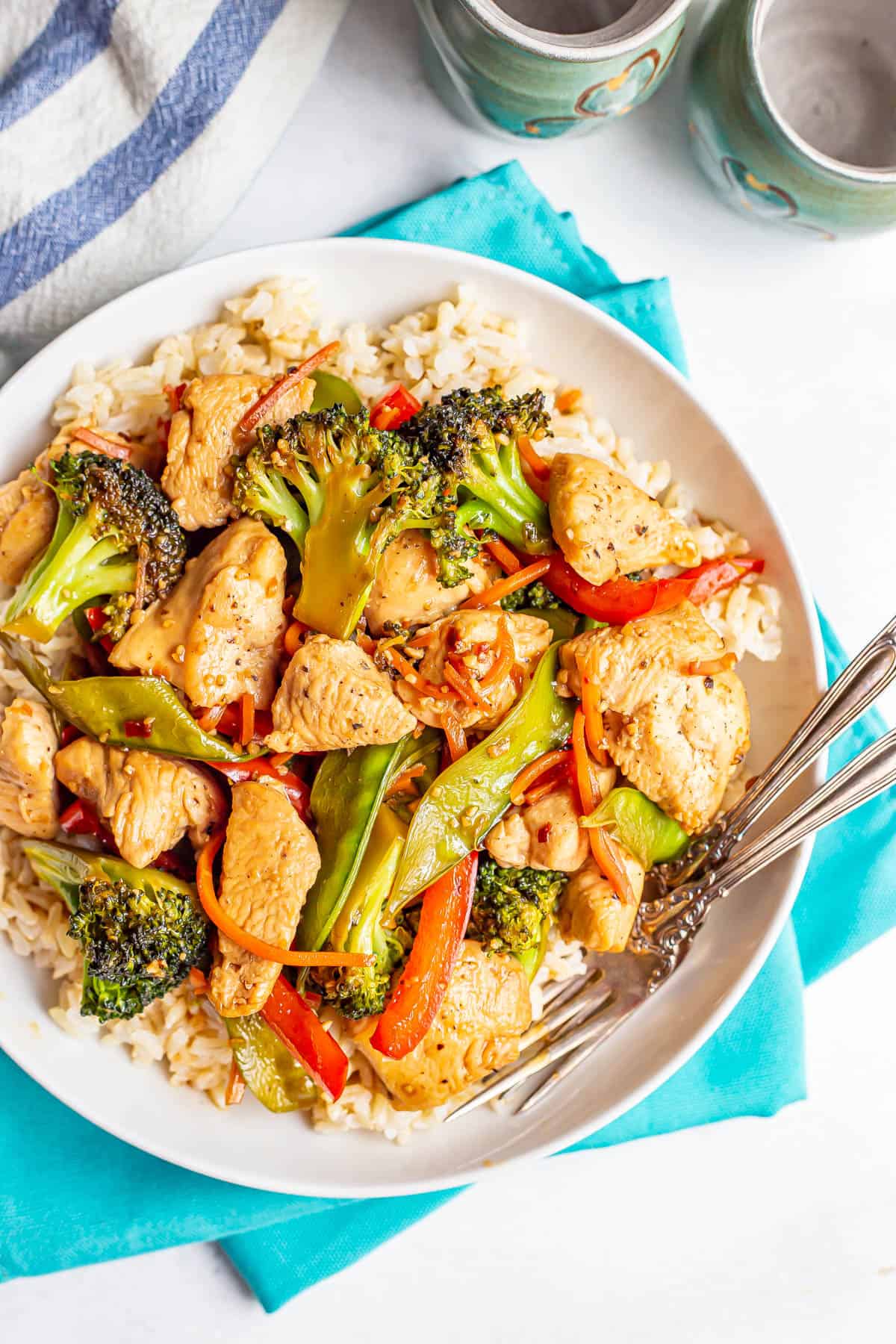 Chicken and vegetable stir fry served over steamed brown rice in a low white bowl with forks resting in it.