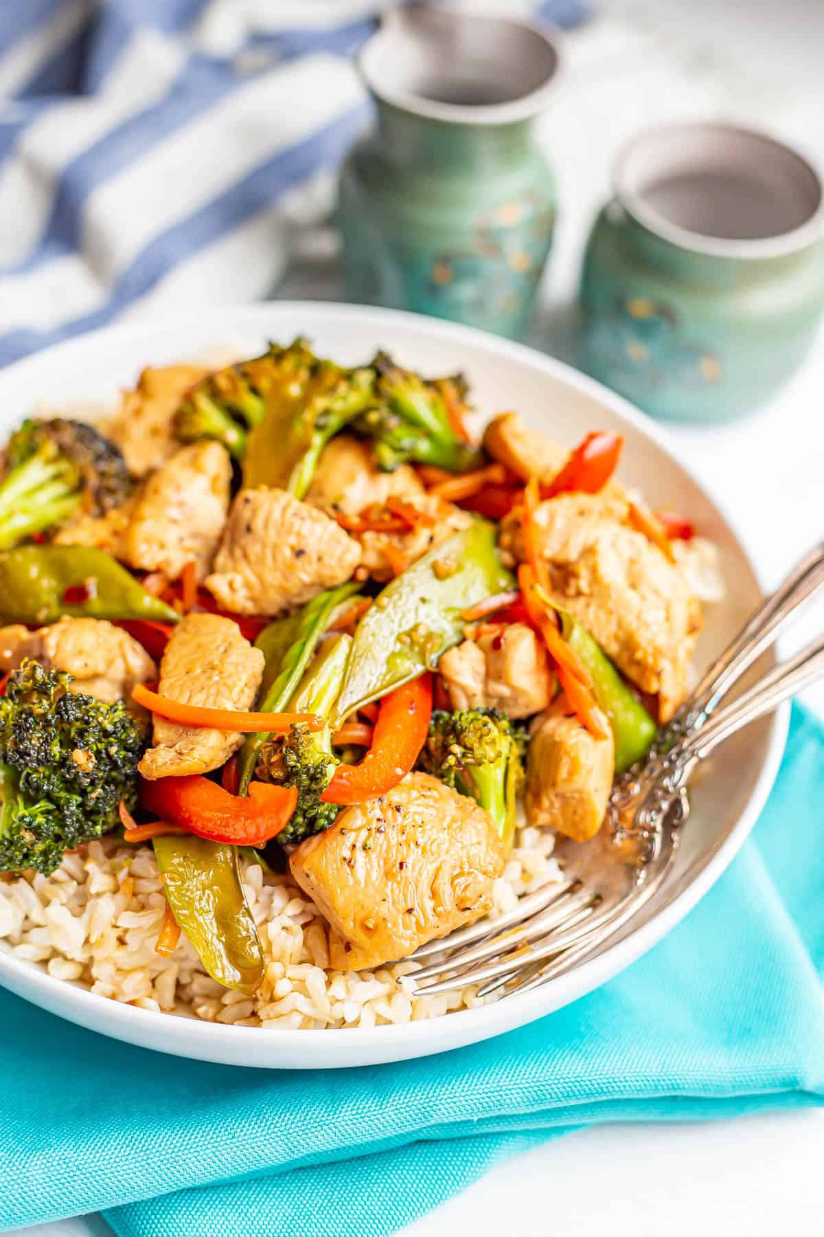 Chicken and vegetable stir fry served over steamed brown rice in a low white bowl with forks resting in it and teal napkins underneath.