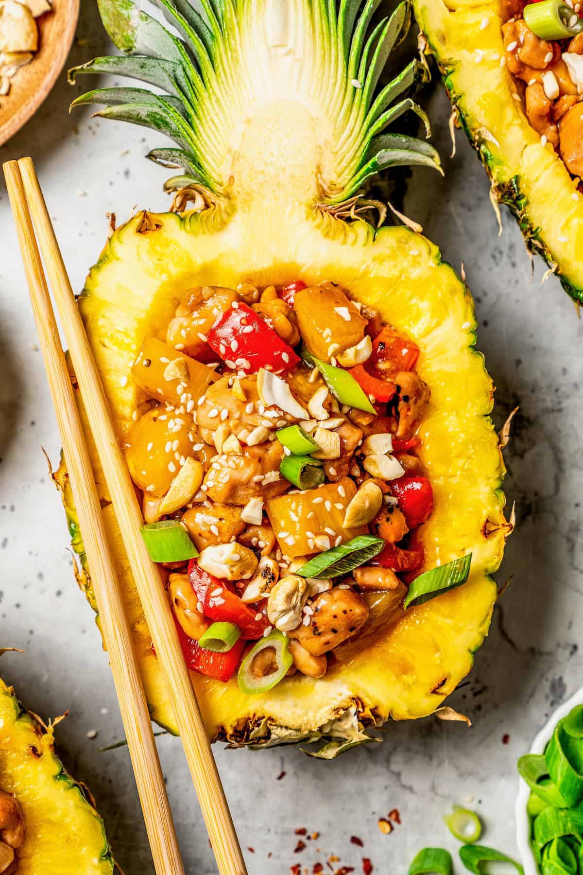 Close up of a chicken and vegetable mix served in a hulled out half of a pineapple with chopsticks resting on the side.
