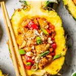 Close up of a chicken and vegetable mix served in a hulled out half of a pineapple with chopsticks resting on the side and a text overlay on the photo.