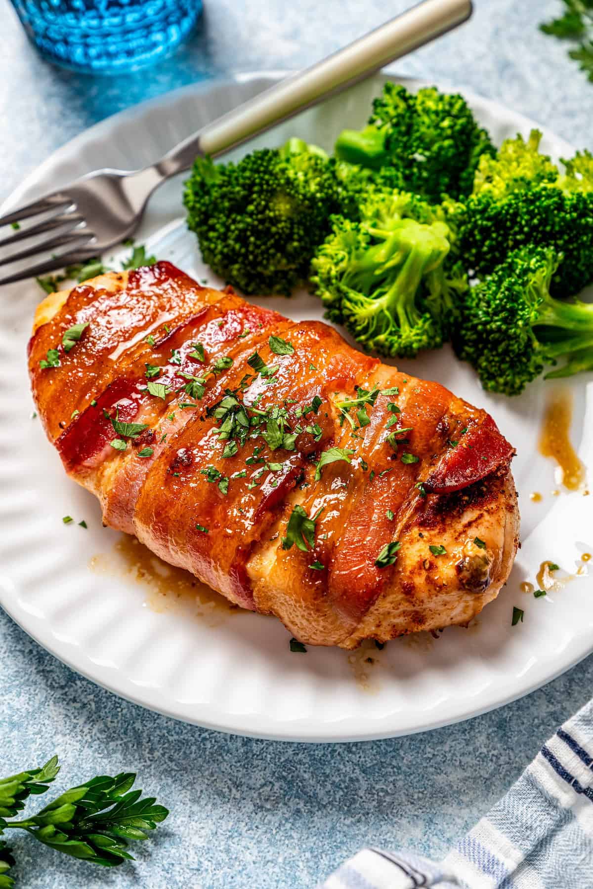 A bacon wrapped chicken breast served on a white plate alongside broccoli.