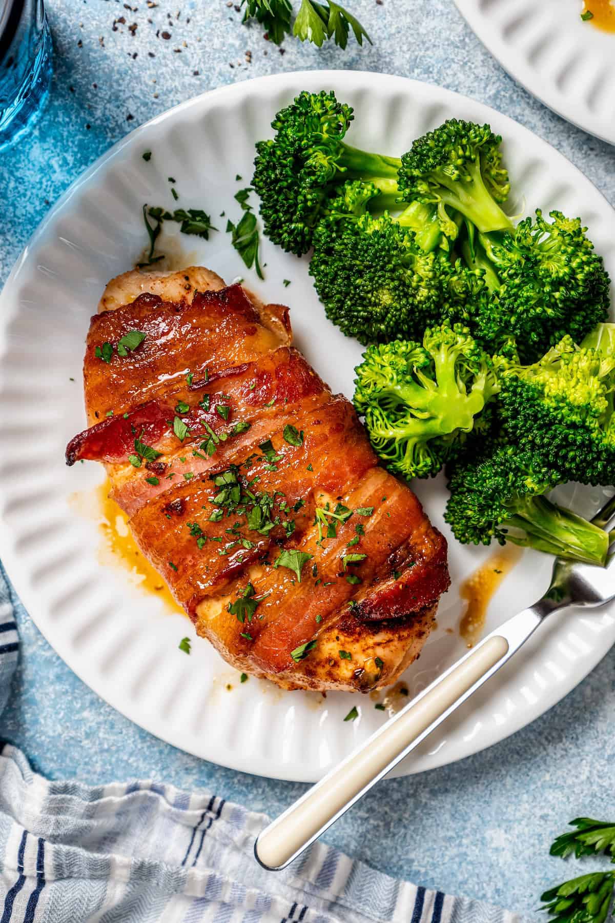 A bacon wrapped chicken breast served on a white plate alongside broccoli with a fork to the side.