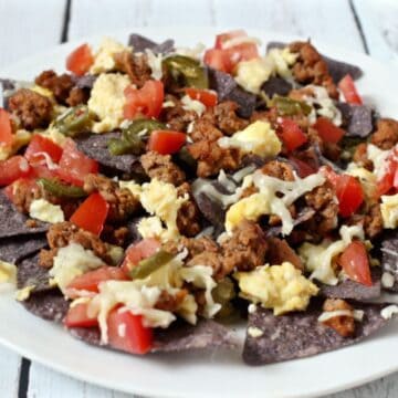 A white plate loaded with breakfast nachos with scrambled eggs, breakfast sausage and toppings on blue chips.