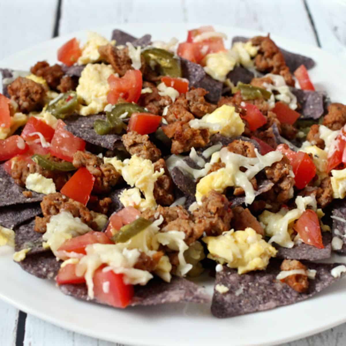 A white plate loaded with breakfast nachos with scrambled eggs, breakfast sausage and toppings on blue chips.