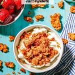 Greek yogurt topped with granola in a small white bowl with granola clusters scattered around and a bowl of strawberries to the side and a text overlay on the photo.