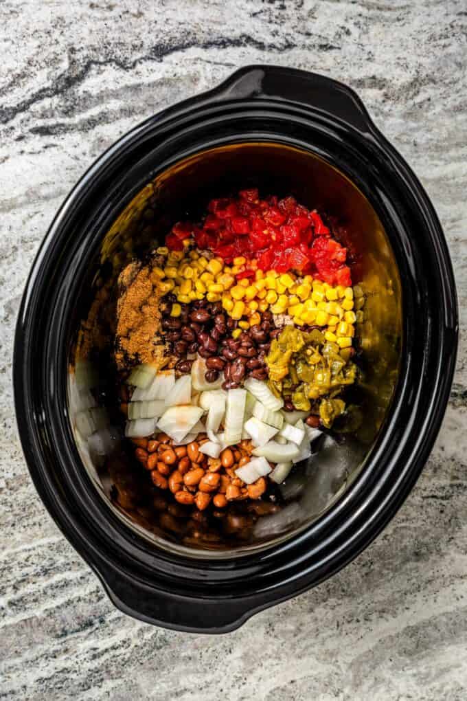 A black slow cooker with tomatoes, corn, beans, chilies, onions and seasonings before being mixed.