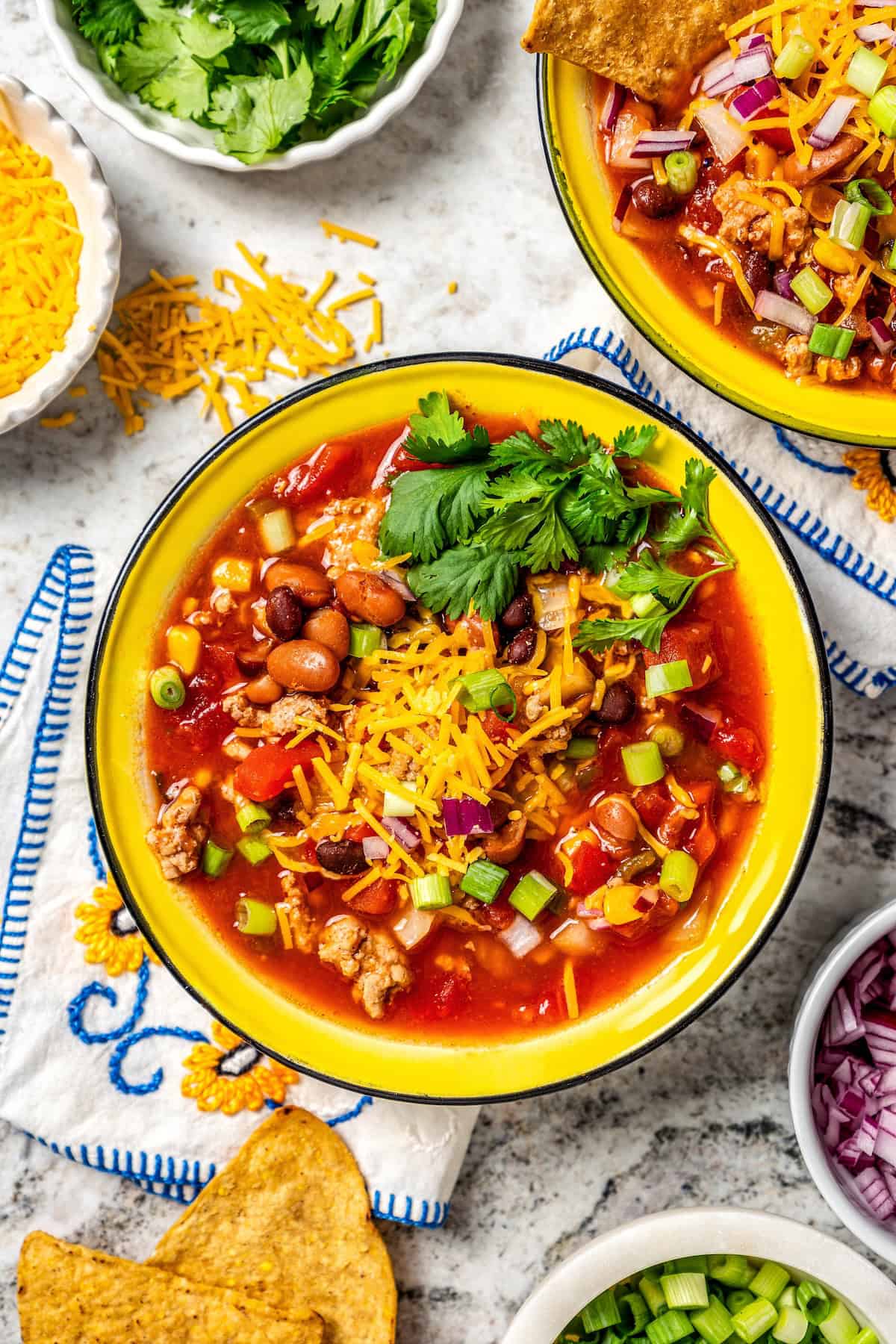 A bowl full of taco soup with cheese and cilantro on top and additional toppings in bowls to the side.