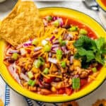 Crock pot taco soup served in a yellow bowl with green onions, red onions, cheese, cilantro and tortilla chips as toppings and a text overlay on the photo.
