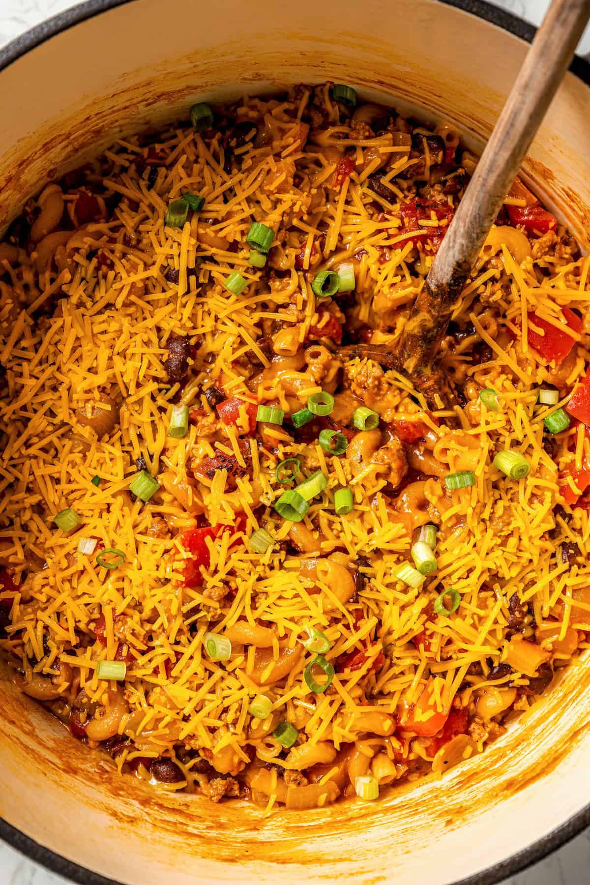 A wooden spoon resting in a large pot of cheesy taco pasta.