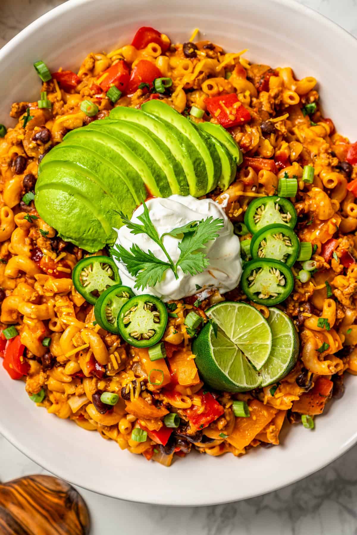 A large serving bowl of cheesy taco pasta with avocado, sour cream, jalapenos and lime wedges on top.