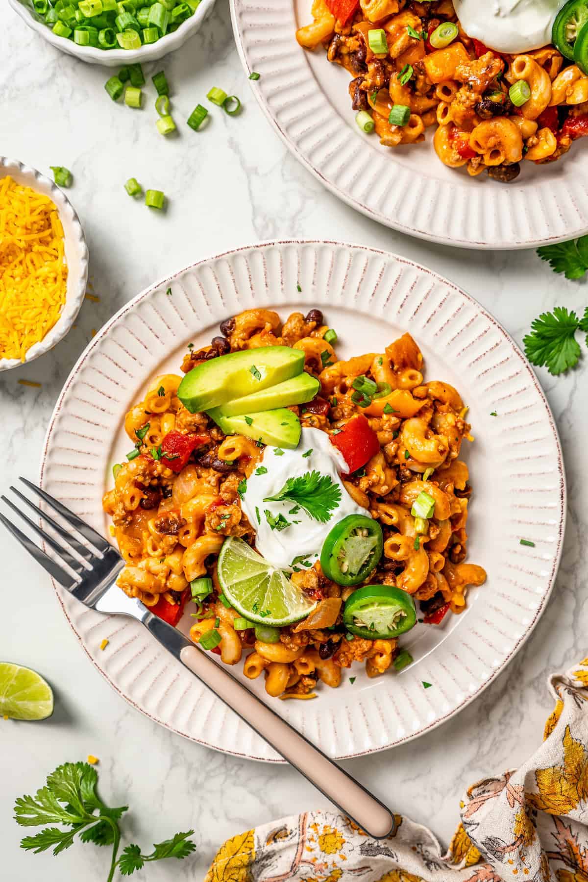 Two plates with taco pasta and avocado, sour cream, jalapenos and lime as toppings with a fork to the side of one plate.