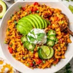 A large serving bowl of cheesy taco pasta with avocado, sour cream, jalapenos and lime wedges on top and a wooden spoon resting in the side and a text overlay on the photo.
