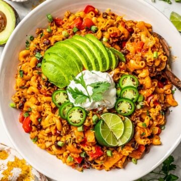 Close up of a large serving bowl of cheesy taco pasta with avocado, sour cream, jalapenos and lime wedges on top and a wooden spoon resting in the side for serving.