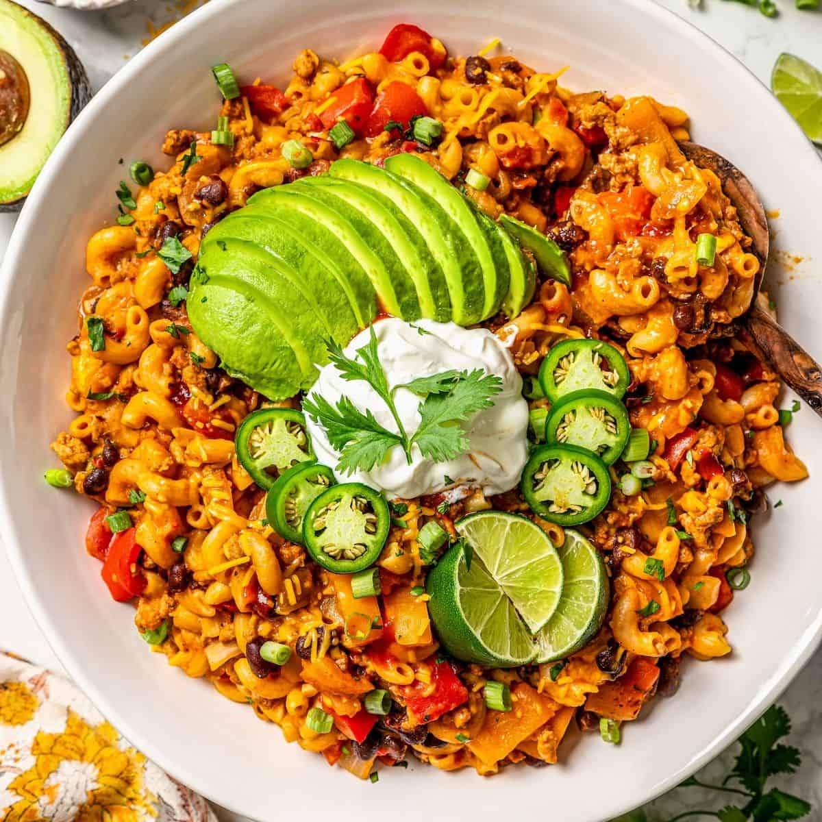 Close up of a large serving bowl of cheesy taco pasta with avocado, sour cream, jalapenos and lime wedges on top and a wooden spoon resting in the side for serving.
