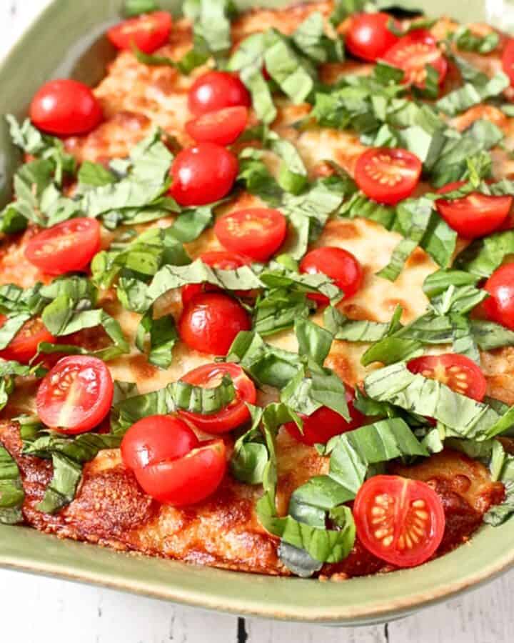 Close up of a green casserole dish with a cheesy baked quinoa topped with sliced cherry tomatoes and fresh basil.