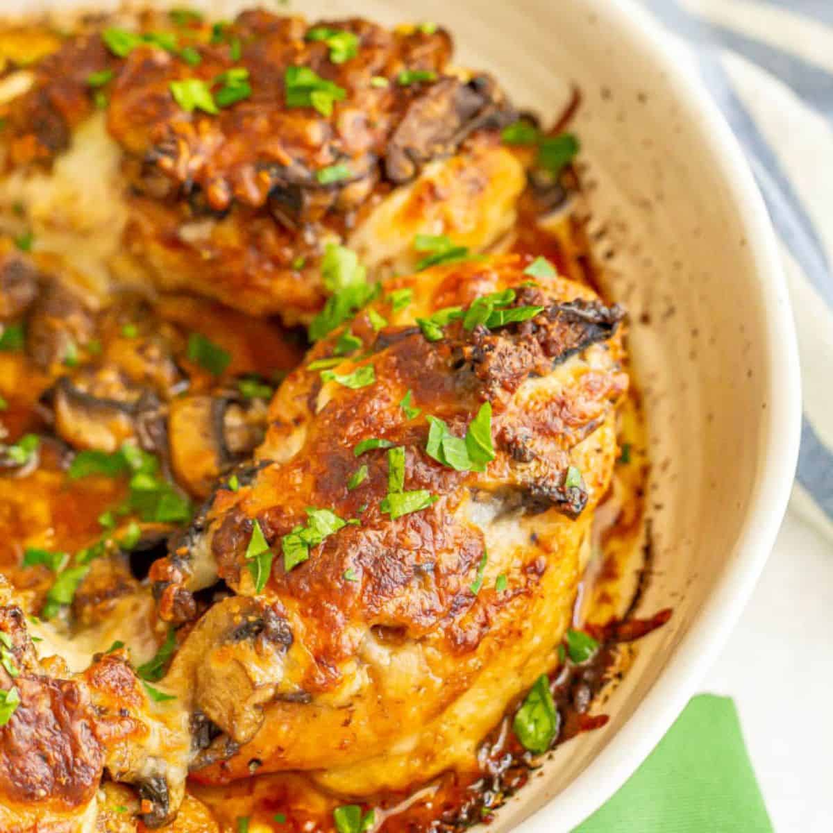 Close up of a white round casserole dish with chicken breasts covered with mushrooms and cheese and topped with parsley.