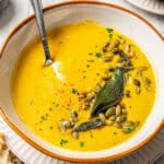 A spoon tucked into a rimmed white low bowl with butternut squash soup topped with pepitas and herbs and a text overlay on the photo.