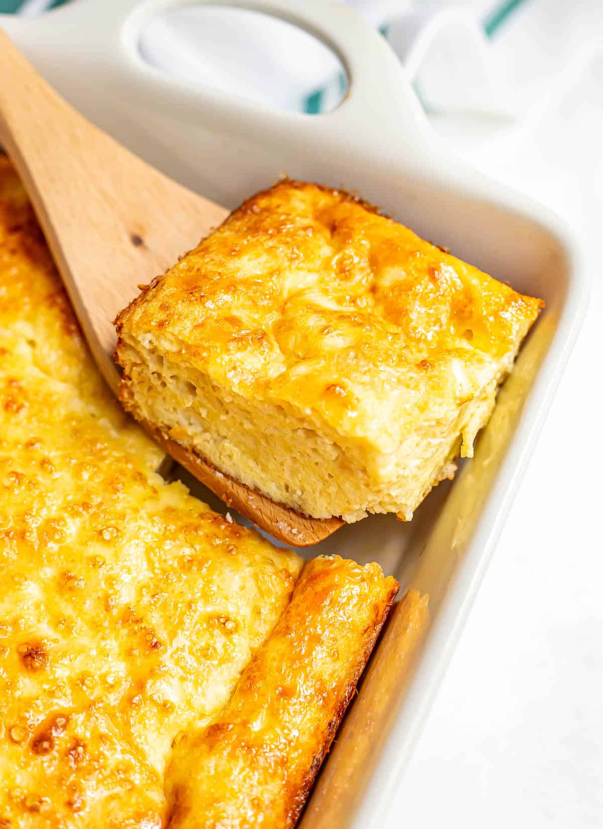 A wooden spatula lifting a square of cheesy baked eggs from a casserole dish.