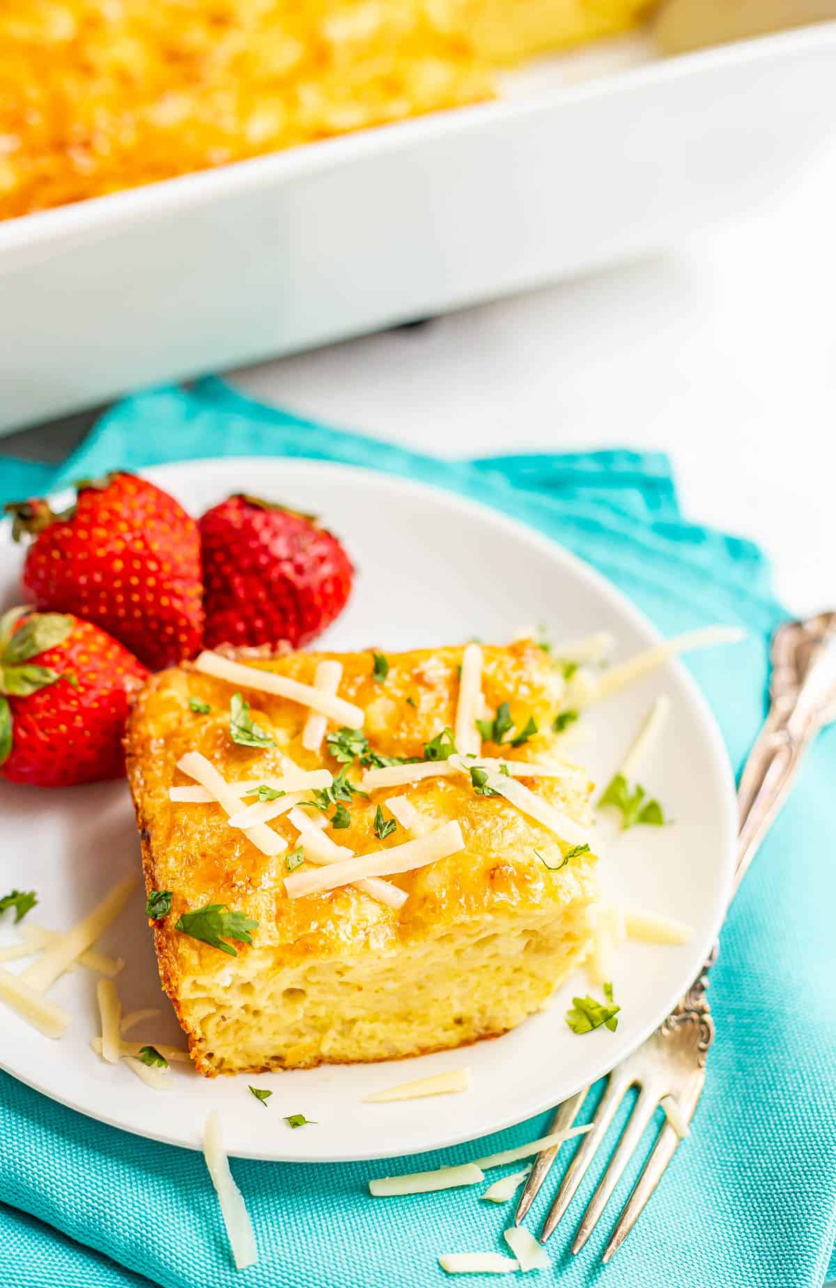 A square of baked eggs topped with cheese and fresh herbs on a white plate alongside some fresh strawberries.