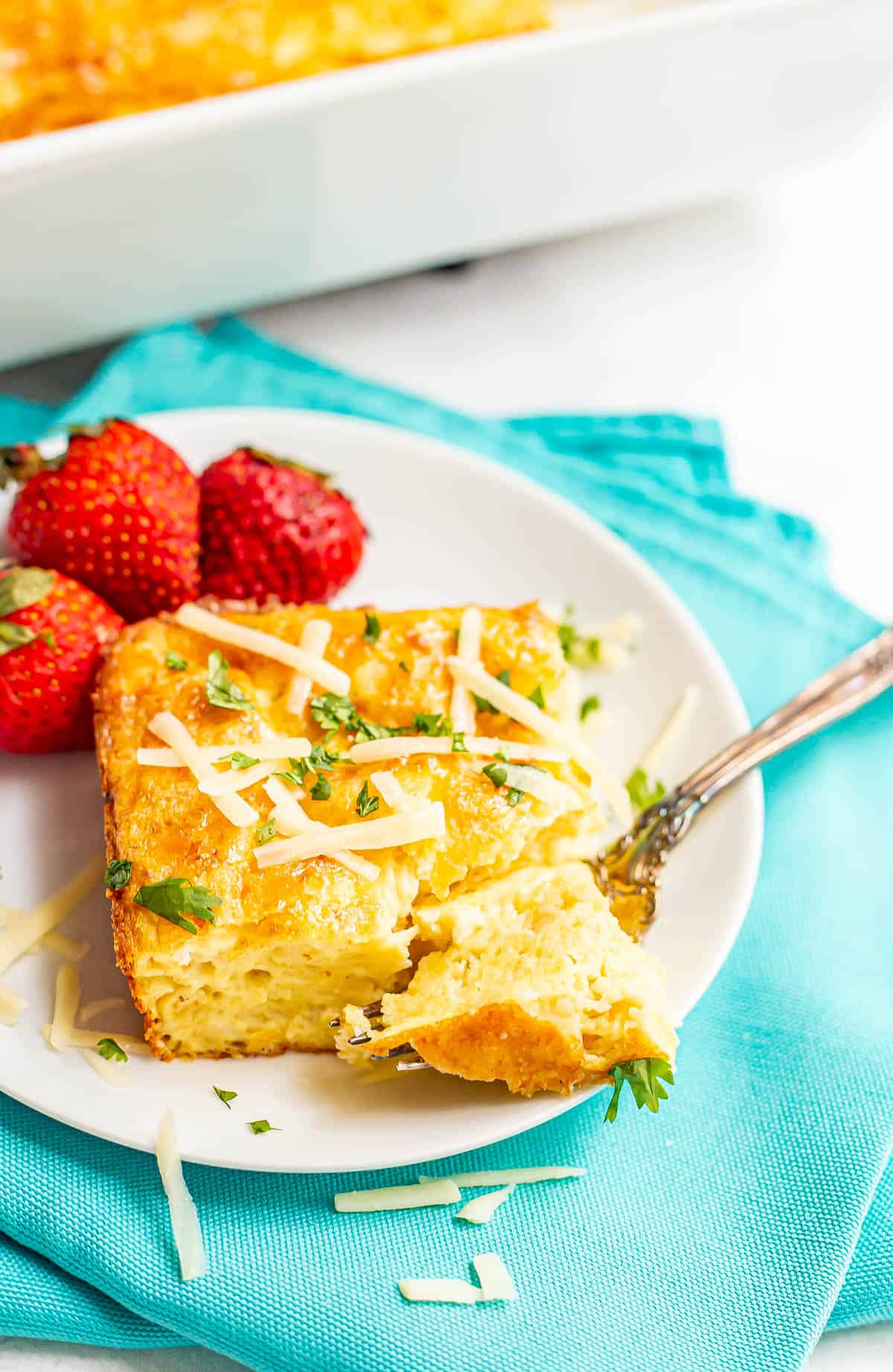 A fork resting into a bite of a square of baked eggs topped with cheese and fresh herbs on a white plate alongside some fresh strawberries.
