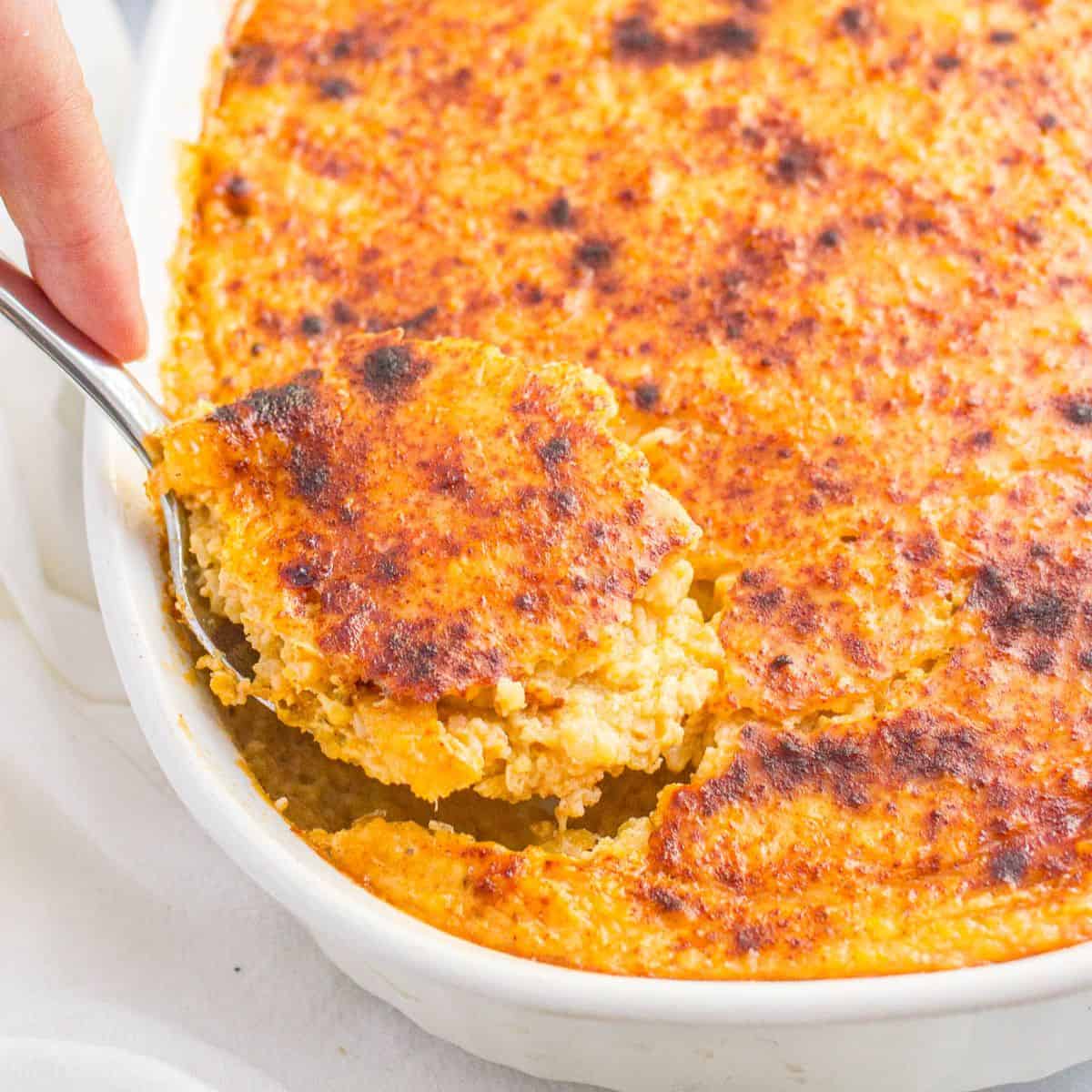 Close up of a hand holding a serving spoon in an oval white casserole dish with a cheesy grits casserole.