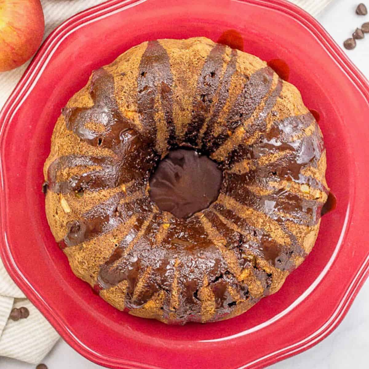 Overhead view of a chocolate chip apple cake drizzled with chocolate sauce and served on a red round platter.
