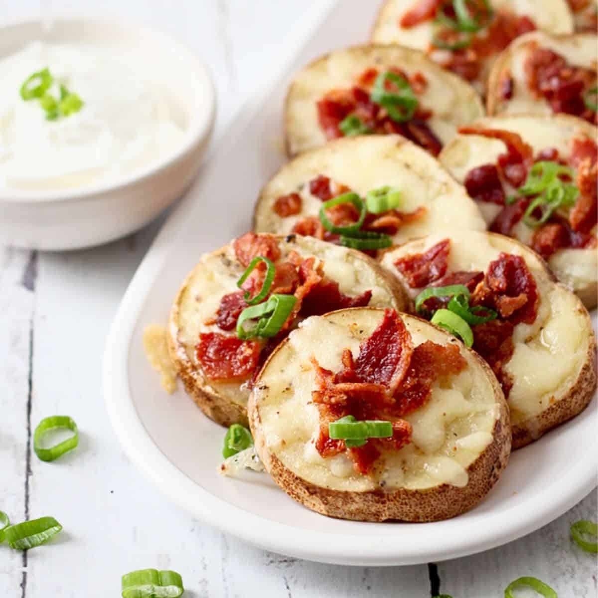 Close up of an oval platter of sliced potato skins with cheese and bacon served alongside a small bowl of sour cream.