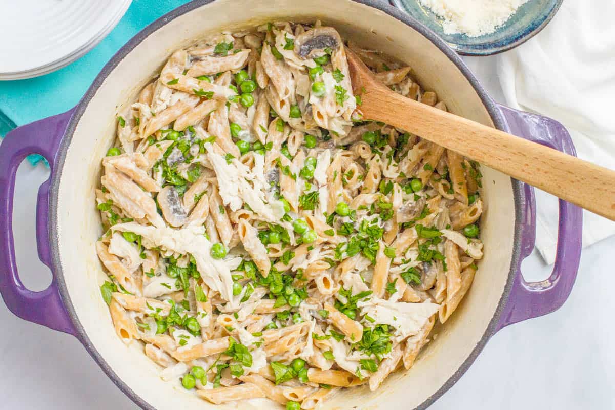 A wooden spoon resting in a large purple dutch oven with a creamy one pot pasta with chicken and mushrooms and peas topped with fresh parsley.