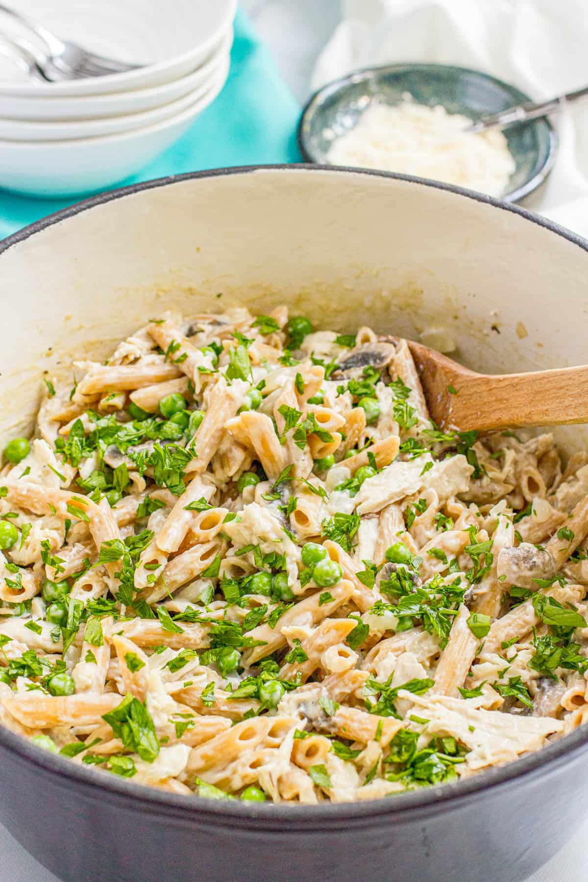 A wooden spoon resting in a large purple dutch oven with a one pot pasta with chicken and mushrooms and peas topped with fresh parsley.