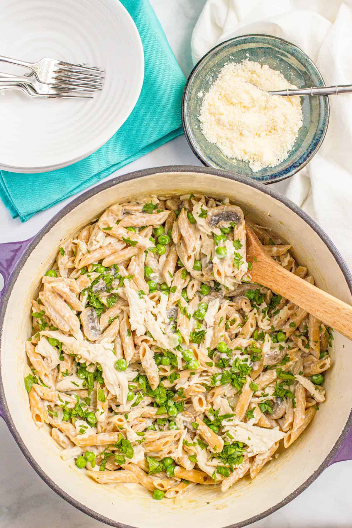 A wooden spoon resting in a large purple dutch oven with a one pot pasta with chicken and mushrooms and peas topped with fresh parsley and a bowl of Parmesan cheese to the side.