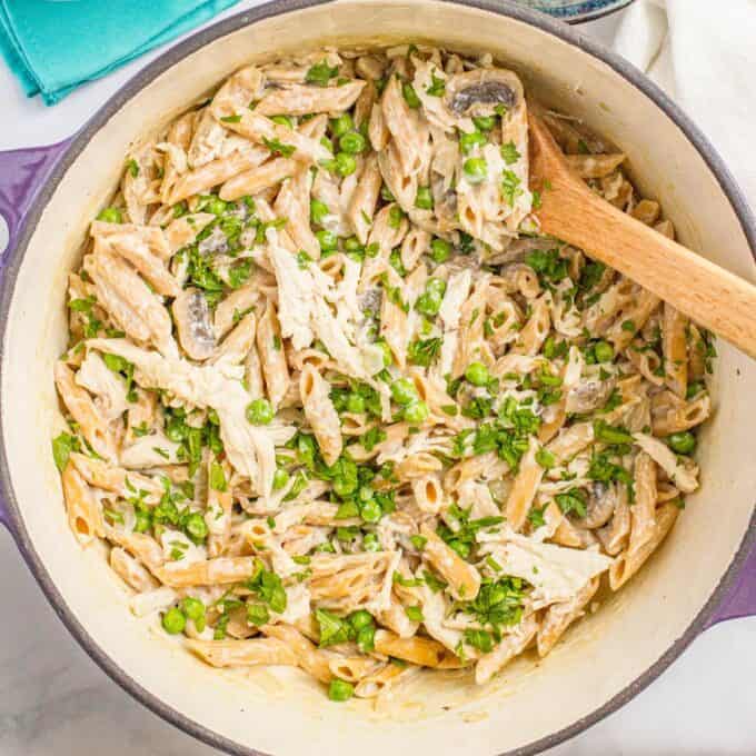 Close up of a wooden spoon resting in a large purple dutch oven with a one pot pasta with chicken and mushrooms and peas topped with fresh parsley.