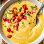 Close up of a spoon resting in a white bowl of potato leek soup with cooked bacon, hot sauce and fresh herbs on top with a text overlay on the photo.