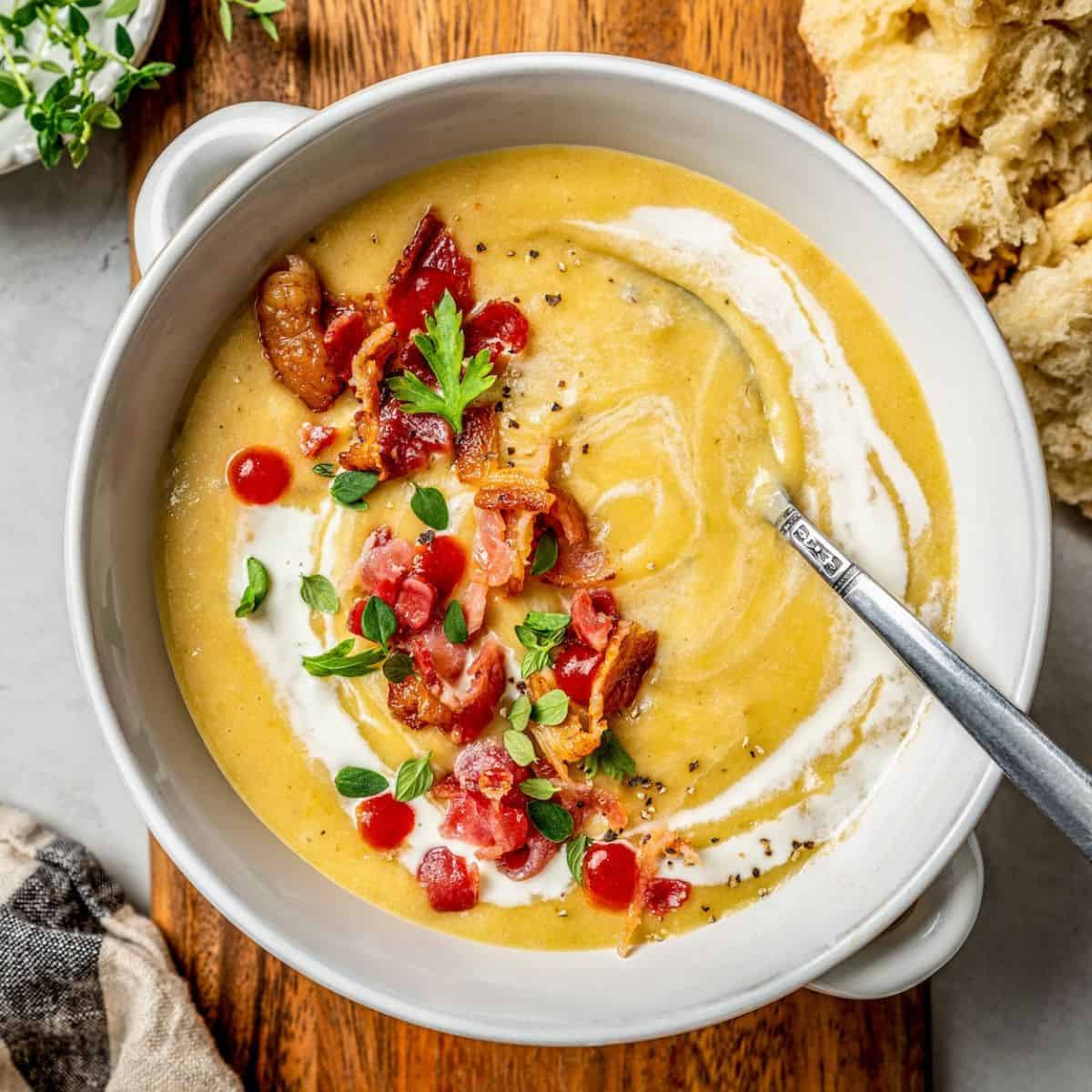 Close up of a spoon resting in a white soup bowl of potato leek soup with bacon and herbs and a swirl of heavy cream, set on a wooden cutting board.
