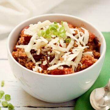 Close up of a white bowl with a vegetarian quinoa chili with beans topped with shredded white cheddar cheese and sliced green onions.