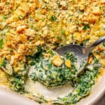 Close up of a spoon resting in a baking dish of creamed spinach casserole with melted cheese and golden brown buttery cracker crumbs on top with a text overlay on the photo.