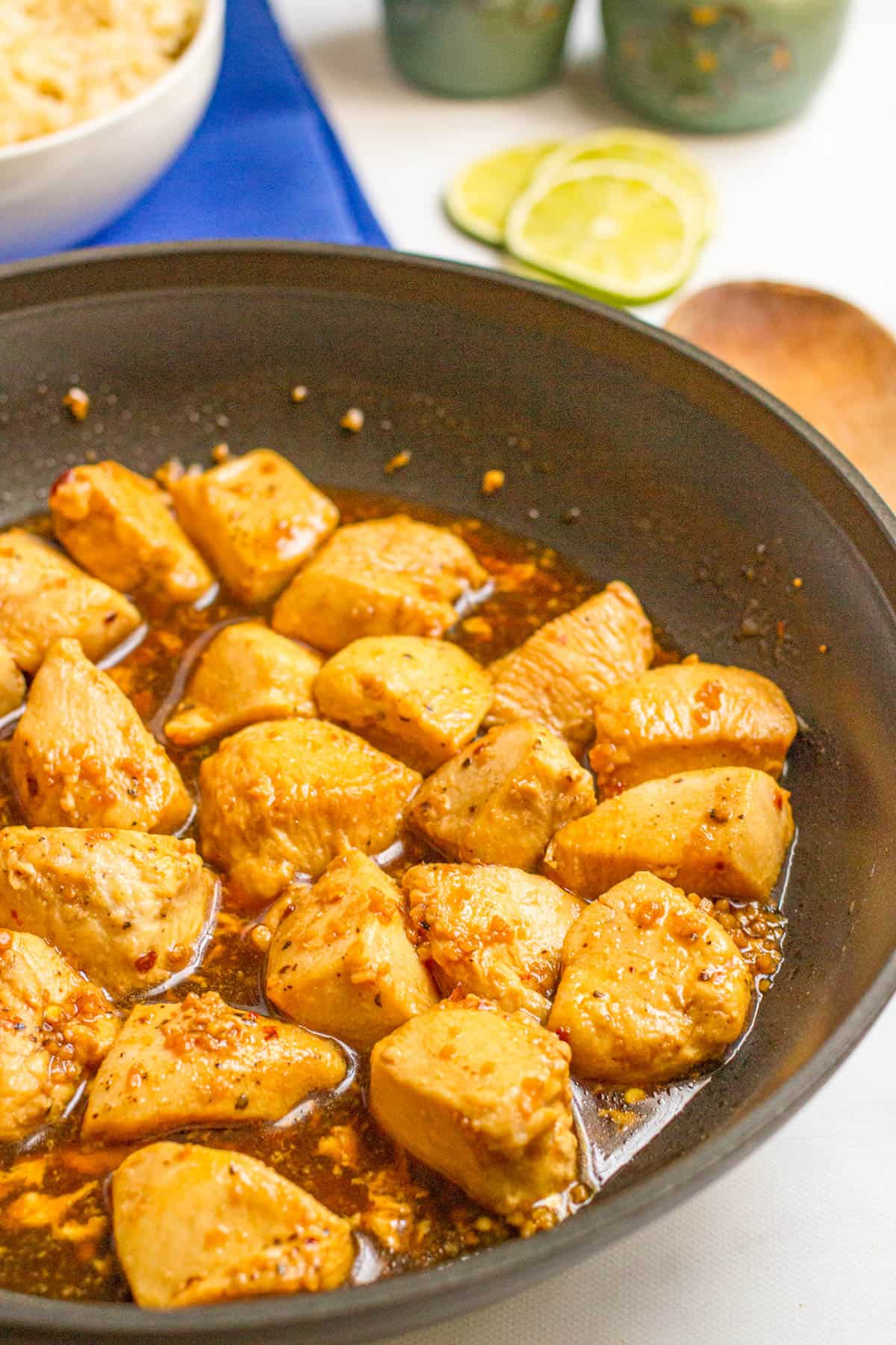 Close up of a large dark skillet with cubed chicken pieces in a honey garlic sauce, with rice in a bowl in the background.